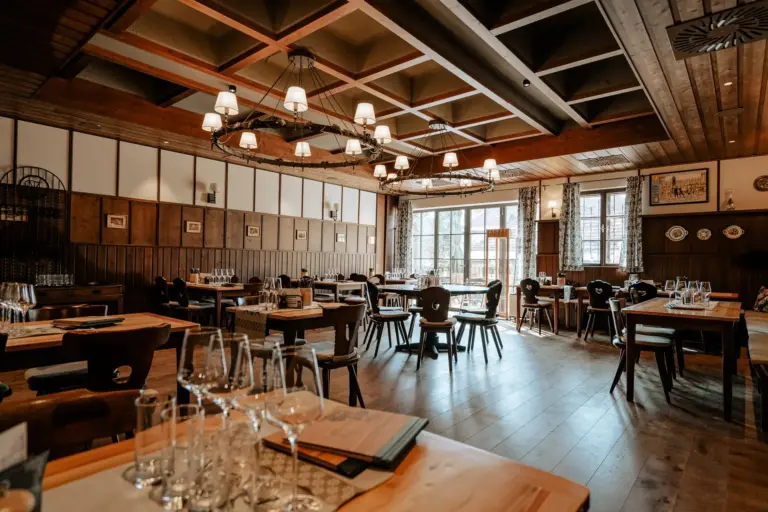 Geräumiger, rustikaler Speisesaal mit Holzböden, Tischen und Stühlen. Von einer Kassettendecke hängen Kronleuchter. Das Sonnenlicht strömt durch große Fenster herein und erhellt die ordentlich gedeckten Tische mit Gläsern und Speisekarten.