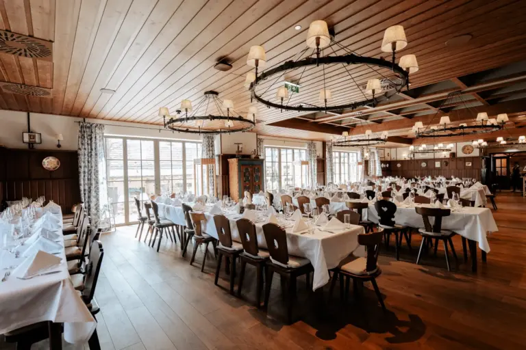 Ein geräumiger, eleganter Speisesaal mit Holzfußboden und -decke, der für eine Veranstaltung mit Tischreihen aus weißen Tüchern, gefalteten Servietten und Weingläsern gedeckt ist und von großen runden Kronleuchtern beleuchtet wird.