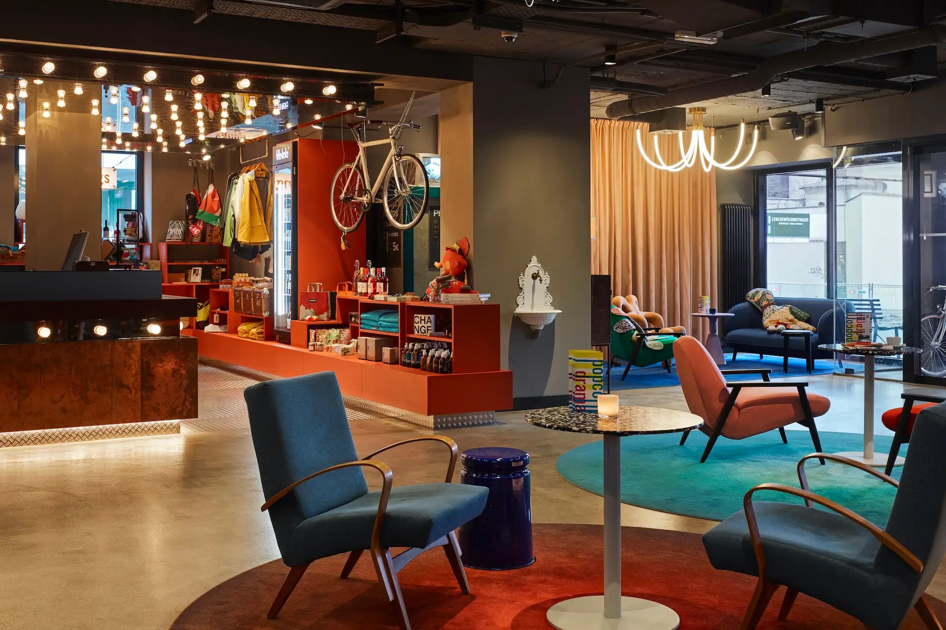 Moderne Hotel-Lobby-Lounge mit blauen und rosa Sesseln, runden Teppichen und einem Fahrrad, das an einer orangefarbenen Wand hinter dem Empfangstresen befestigt ist.