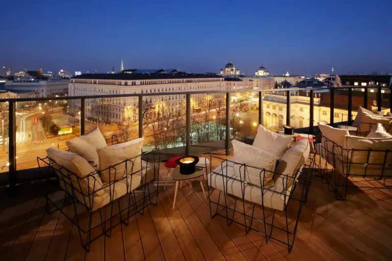 Dachterrasse mit Lounge-Sitzgelegenheiten und Blick auf eine Stadtstraße und historische Gebäude in der Dämmerung.