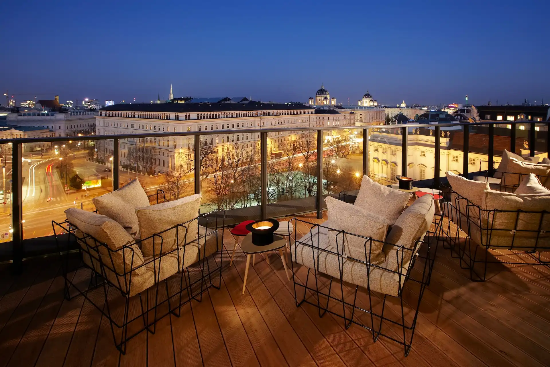 Dachterrasse mit Lounge-Sitzgelegenheiten und Blick auf eine Stadtstraße und historische Gebäude in der Dämmerung.