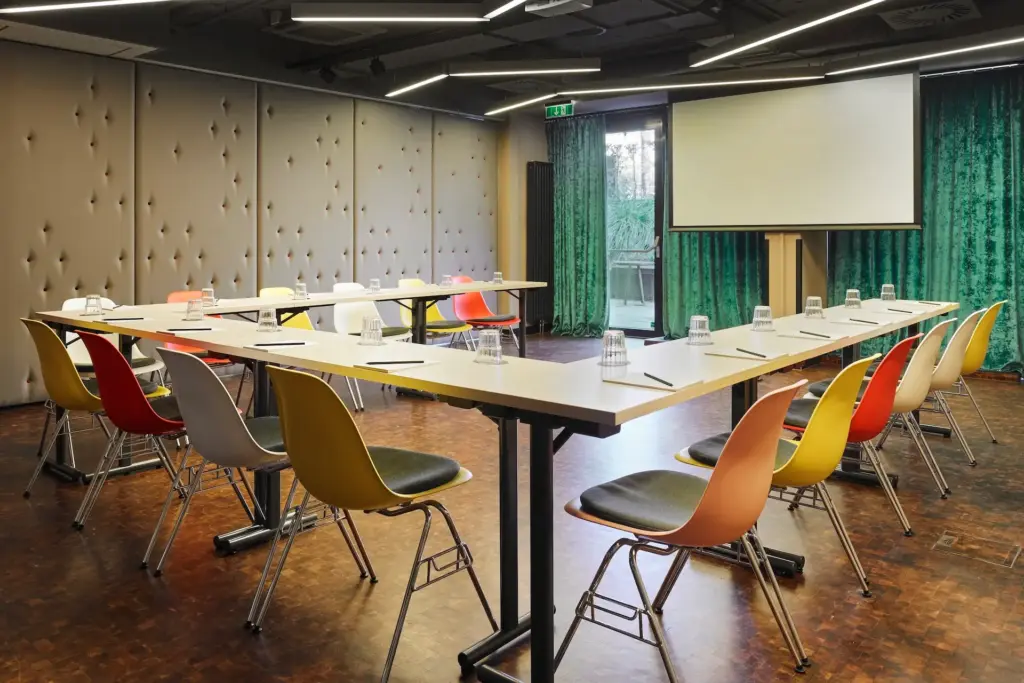 Ein U-förmiger Konferenztisch, umgeben von bunten Stühlen, in einem Besprechungsraum mit einer Projektionswand.