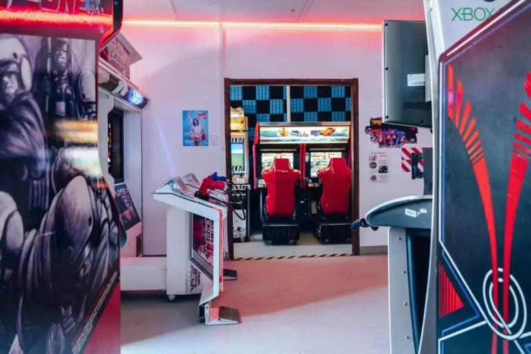 Eine Spielhalle mit verschiedenen Spielautomaten, darunter ein Paar rote Rennsitze und Bildschirme in der Ferne, umgeben von bunten Lichtern und Wandkunst.