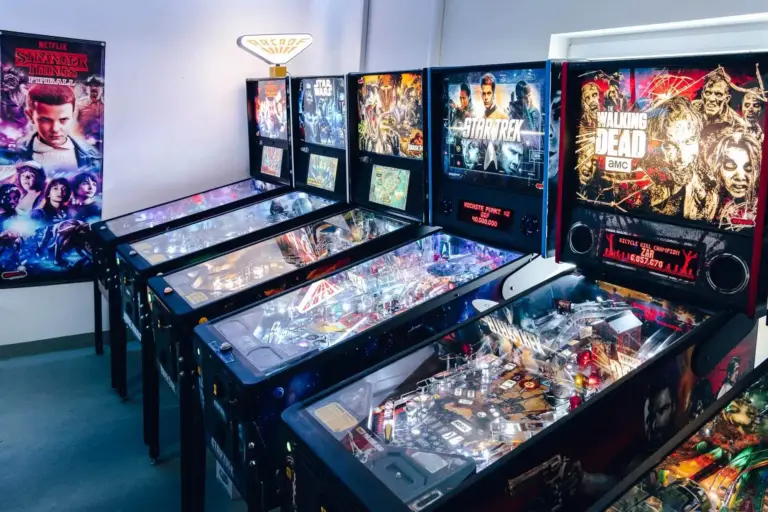 Eine Reihe von Flipperautomaten mit Themen wie Star Trek, The Walking Dead und Stranger Things, aufgereiht in einem hell erleuchteten Spielhallenraum mit einem Stranger Things-Poster an der Wand.