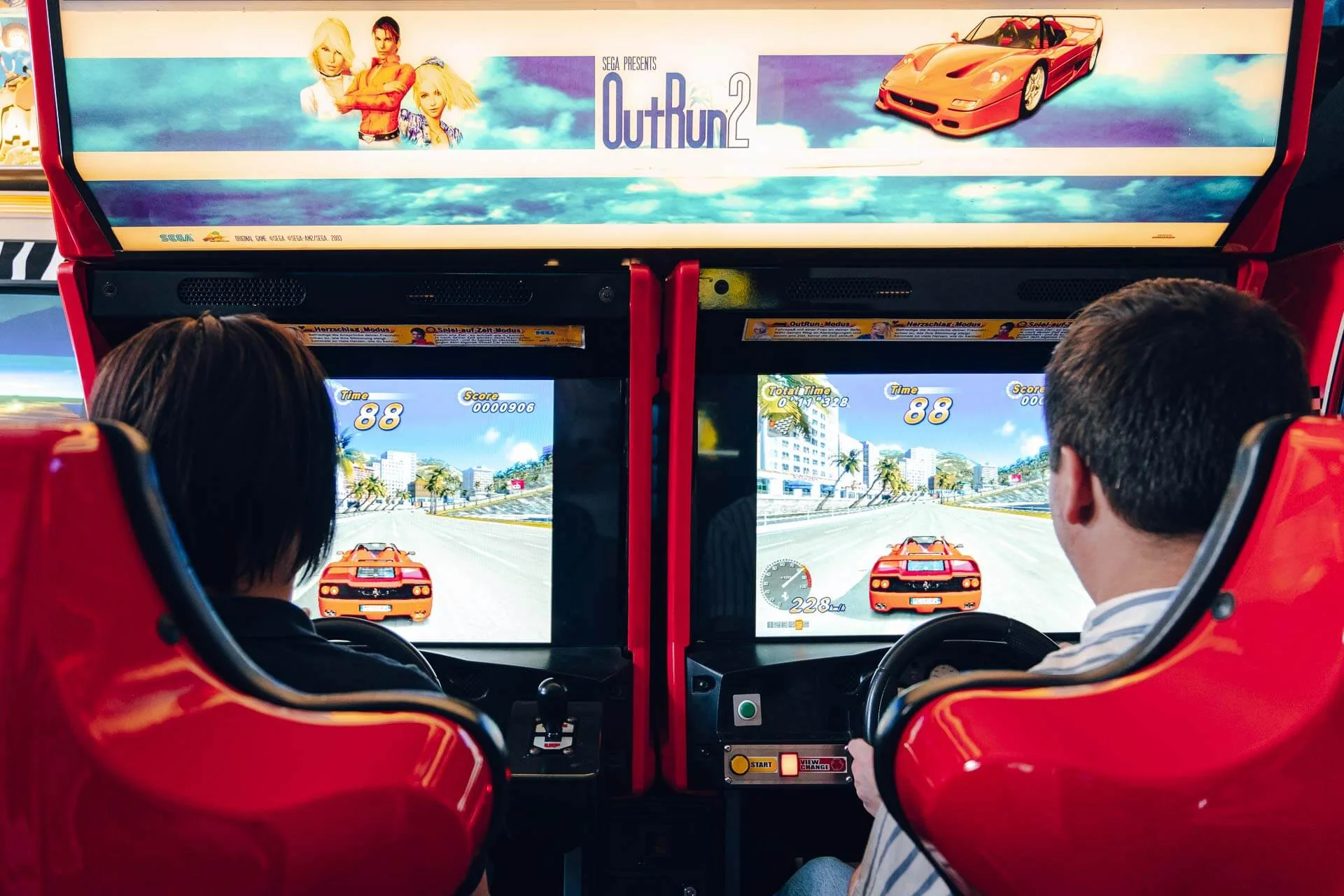 Zwei Personen sitzen nebeneinander und spielen das Arcade-Rennspiel OutRun 2 in roten Fahrsimulatorboxen, wobei jeder einen virtuellen roten Sportwagen auf dem Spielbildschirm steuert.