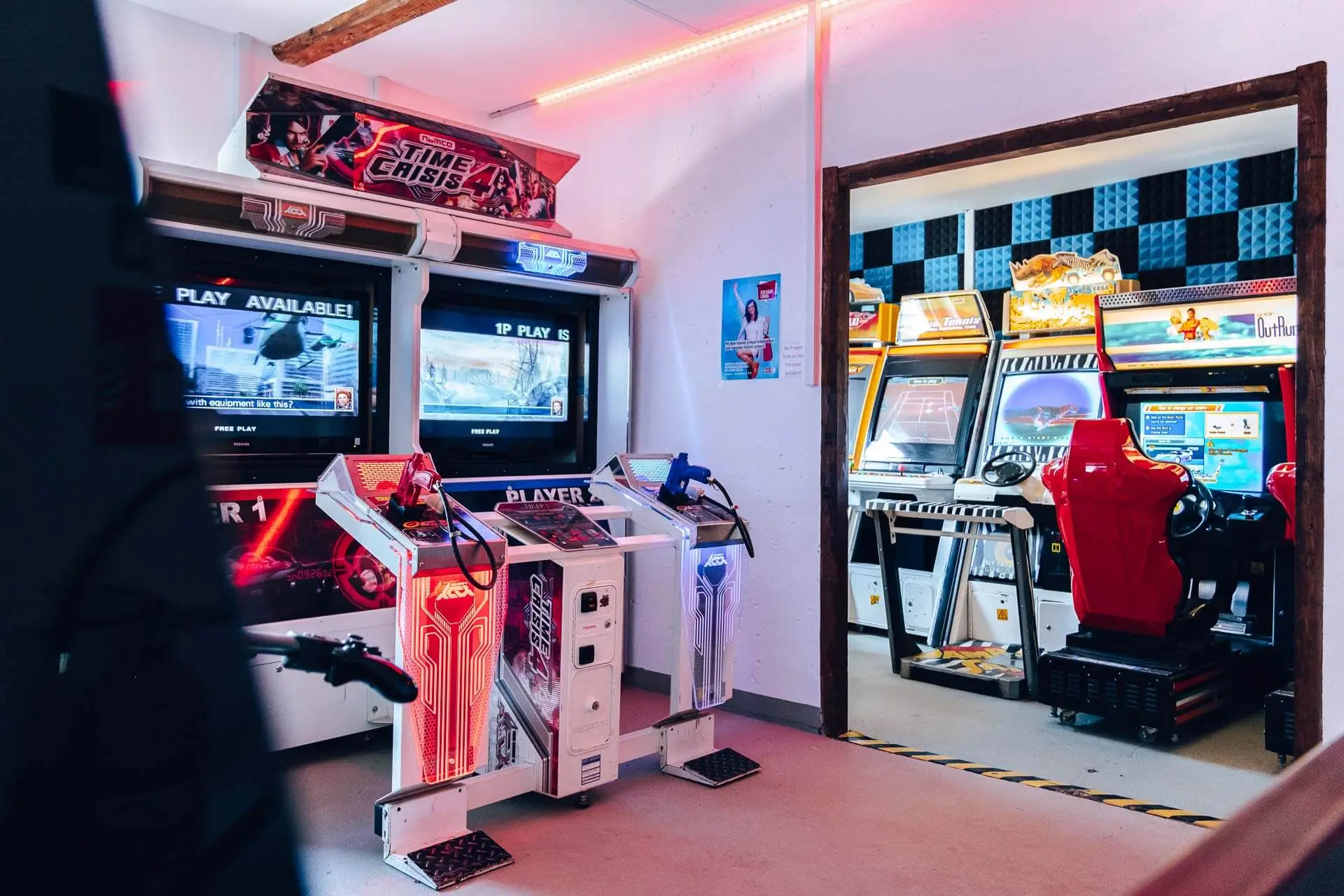 Ein hell erleuchteter Spielhallenraum mit mehreren Retro-Videospielautomaten, darunter zwei Time Crisis-Schießspielschränke im Vordergrund und verschiedene Renn- und Arcade-Spiele, die durch eine offene Tür sichtbar sind.