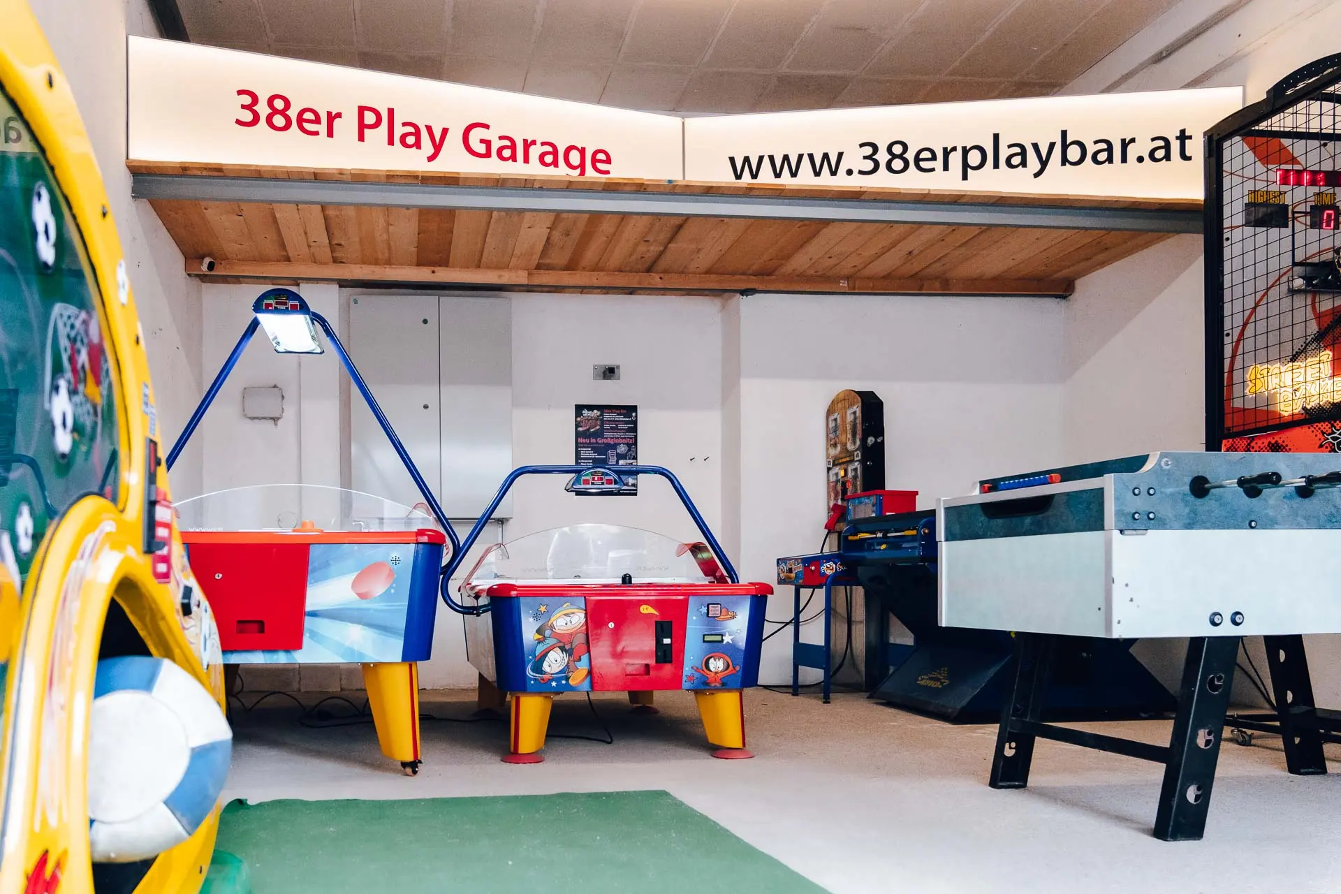 Ein hell erleuchteter Spielhallenraum mit Air-Hockey-Tischen, einem Kickertisch und verschiedenen Spielen. Auf einem großen Schild über dem Raum steht "38er Play Garage" und die Website www.38erplaybar.at.