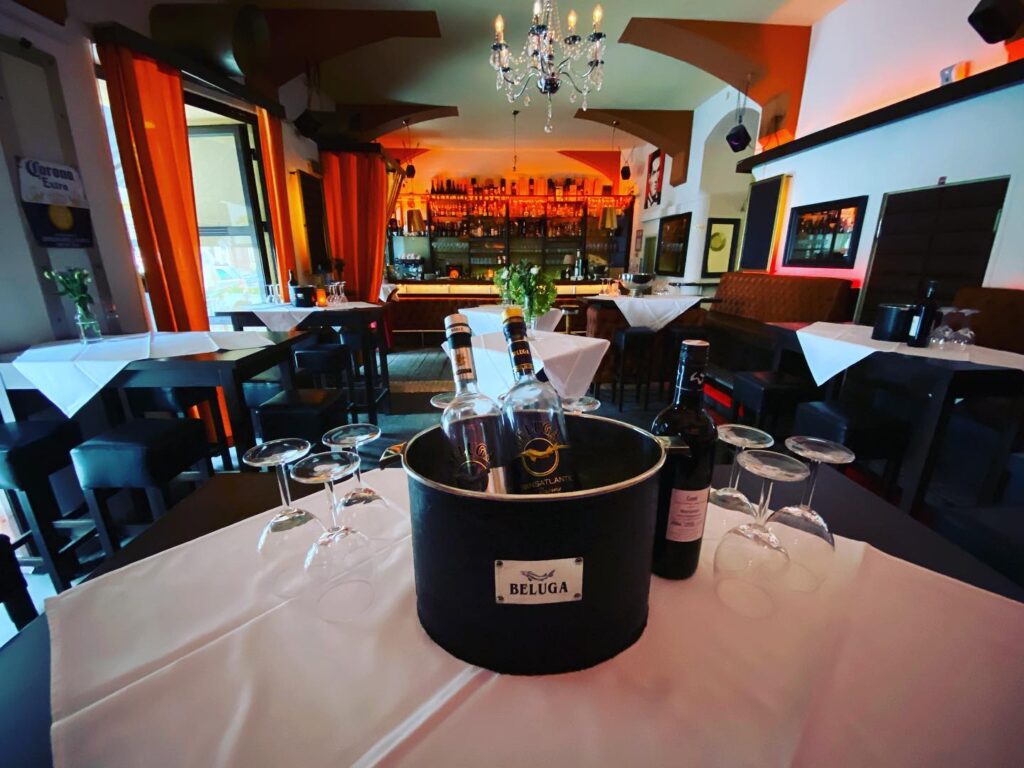 Ein stilvolles Restaurantinterieur mit ordentlich gedeckten Tischen, einem Kronleuchter und warmer Beleuchtung. Im Vordergrund steht ein Eimer mit der Aufschrift Beluga, in dem sich Flaschen befinden, umgeben von sechs leeren Weingläsern auf einer weißen Tischdecke.