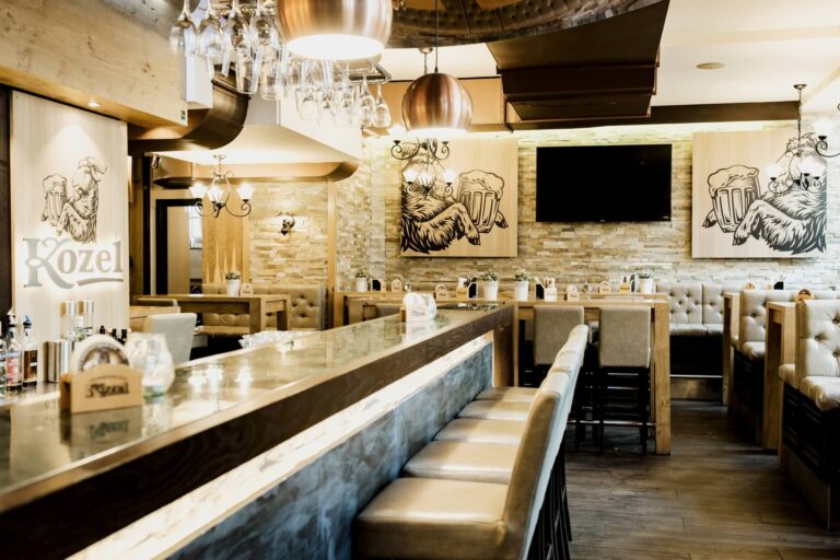 Plutzer Bräu Modernes Barinterieur mit gepolsterten Barhockern, Holztischen und hellbraunen Stühlen. An den Wänden sind Ziegen zu sehen, die Bierkrüge halten, sowie ein großer Fernseher und von der Decke hängende Gläser. Der Raum hat eine gemütliche, gut beleuchtete Atmosphäre.