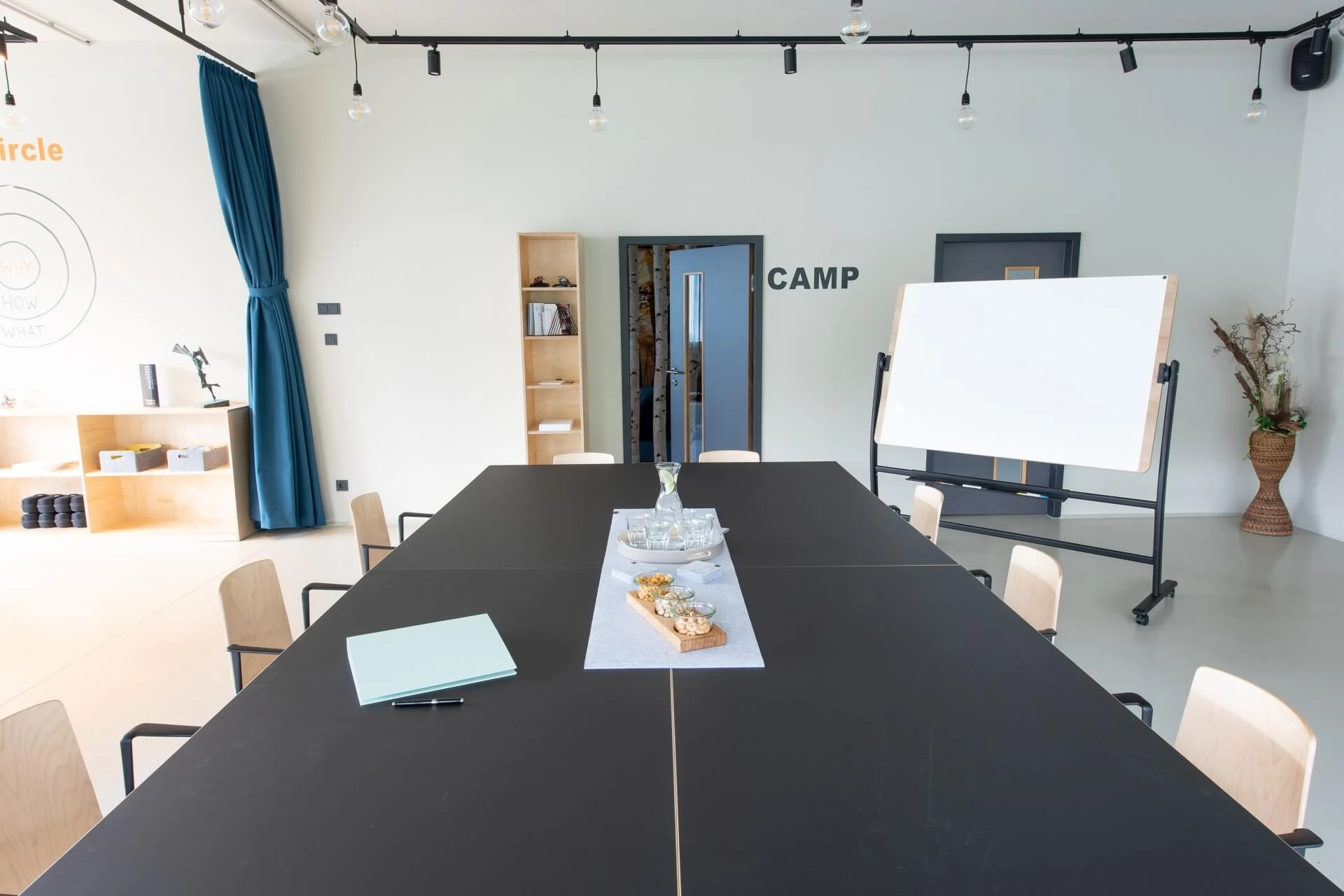 Moderner Konferenzraum mit einem großen schwarzen Tisch, Holzstühlen, einem Whiteboard auf einem Ständer, einem Wasserkrug aus Glas, Snacks in der Mitte und hellen Holzregalen vor weißen Wänden. Blauer Vorhang und dekorative Pflanze im Hintergrund.