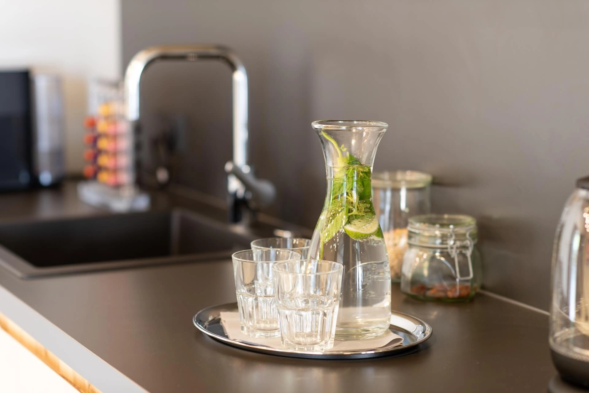 Eine Karaffe Wasser mit Minze und Limette steht zusammen mit drei Gläsern auf einem Metalltablett auf einem modernen Küchentisch in der Nähe einer Spüle. Im Hintergrund stehen Gläser und ein Wasserkocher.