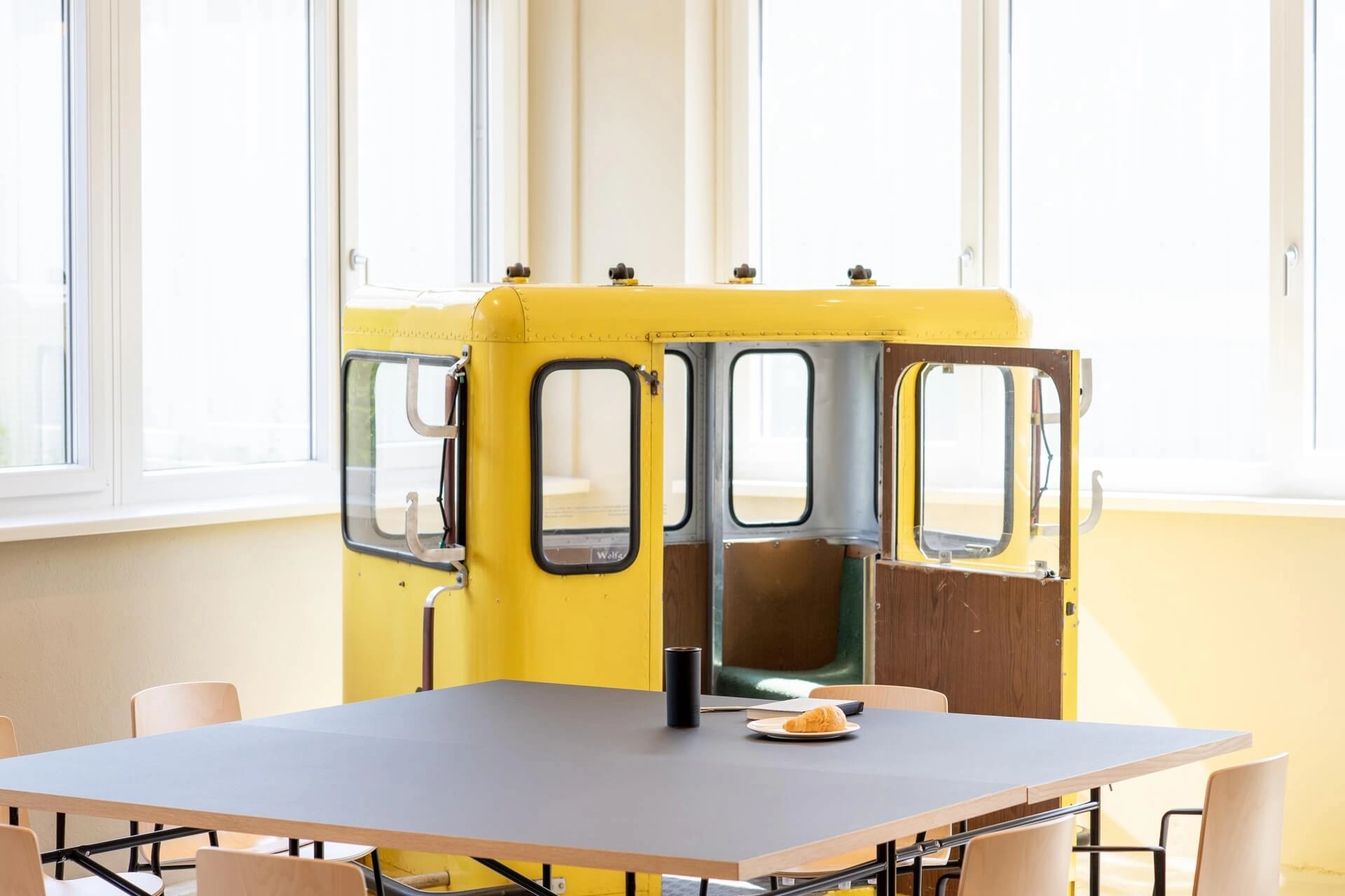 Eine gelbe Kabine, die wie ein Teil eines alten Fahrzeugs oder einer Seilbahn aussieht, steht in einem hellen Raum mit großen Fenstern hinter einem Tisch mit einem Teller, einem Croissant und einer Tasse. Mehrere helle Holzstühle umgeben den Tisch.