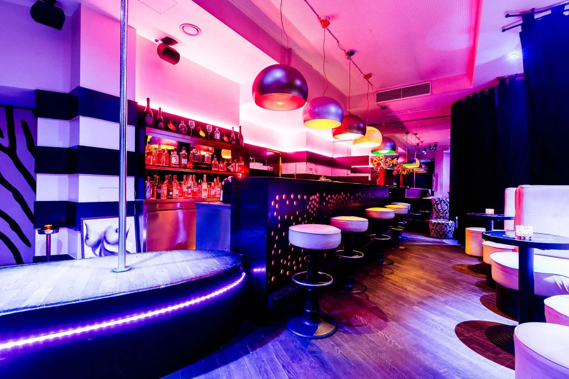 Eine stilvolle Bar mit modernem Dekor, bunter Neonbeleuchtung, einem glänzenden Bartresen, Barhockern und einer kleinen Bühne mit einer Stange. Regale mit Flaschen säumen die Rückwand und sorgen für eine lebhafte, quirlige Atmosphäre im Nachtleben.