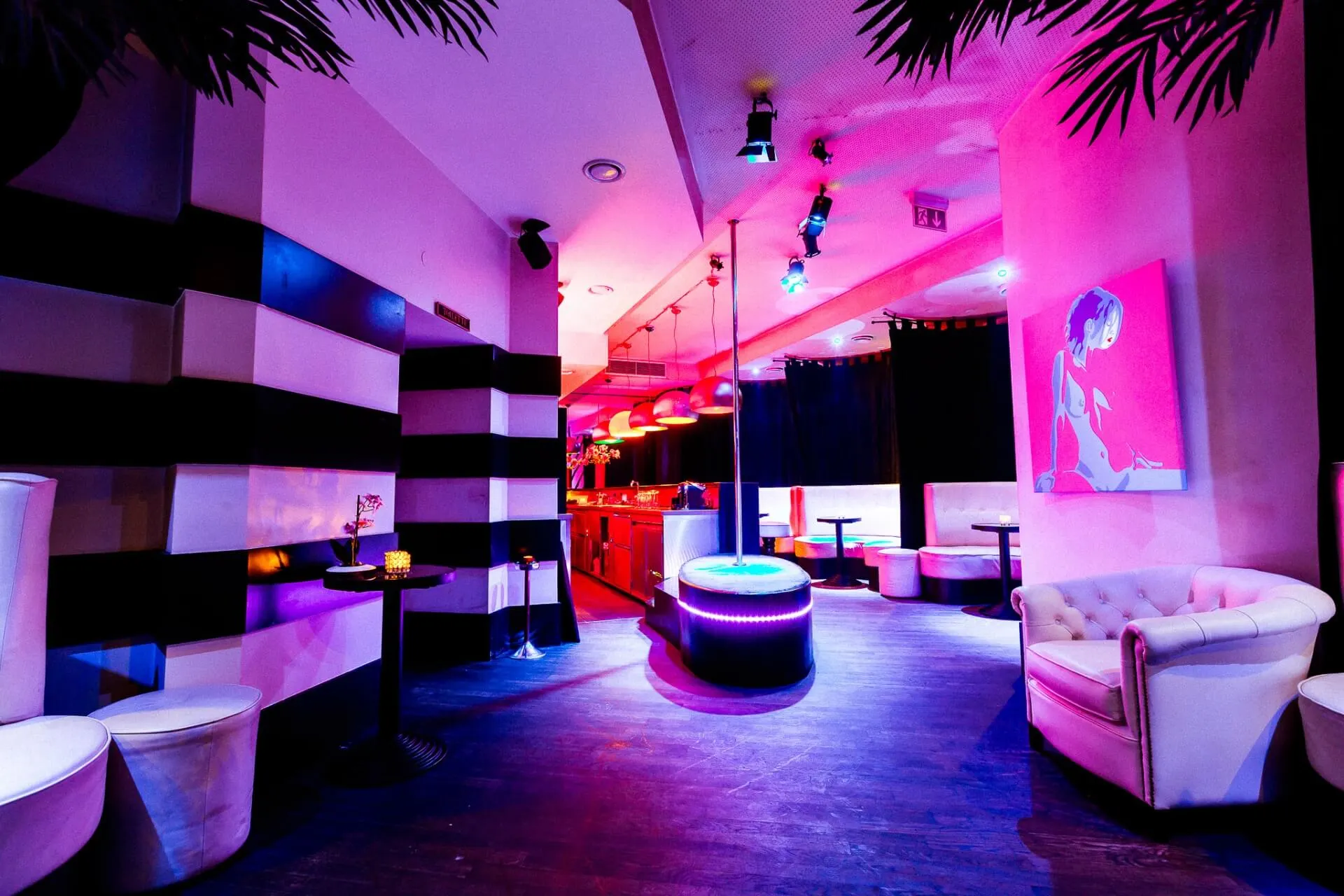Eine moderne Lounge mit rosafarbener und violetter Beleuchtung, weißen Sofas, schwarz-weiß gestreiften Wänden, einem runden Hocker und einer Bar im Hintergrund. Ein buntes Porträt hängt an der Wand.