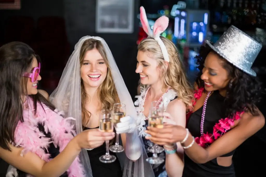 Vier Frauen feiern auf einer After-Wedding-Party, eine als Braut mit Schleier gekleidet, die anderen mit festlichen Accessoires wie Hasenohren, einem silbernen Hut und rosa Boas - alle lächeln und stoßen mit Champagnergläsern an.