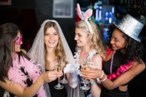 Vier Frauen feiern auf einer After-Wedding-Party, eine als Braut mit Schleier gekleidet, die anderen mit festlichen Accessoires wie Hasenohren, einem silbernen Hut und rosa Boas - alle lächeln und stoßen mit Champagnergläsern an.