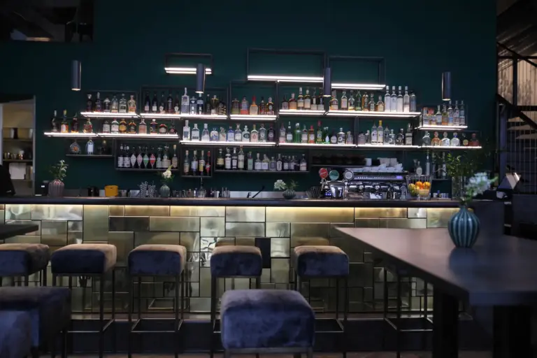 Eine moderne Bar mit hohen Samtstühlen, einem langen Tresen und Regalen mit Schnapsflaschen und Gläsern vor einer dunkelgrünen Wand, die von Deckenlampen sanft beleuchtet wird. Eine Vase mit Blumen steht auf einem Tisch in der Nähe.