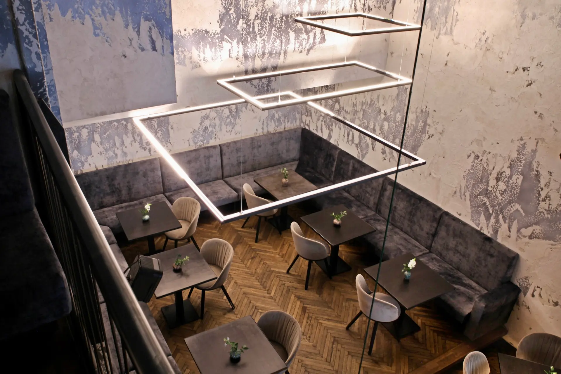 Modernes Restaurantinterieur mit quadratischen Pendelleuchten, Samtkabinen an strukturierten Wänden, kleinen quadratischen Tischen und gepolsterten Stühlen, die auf einem Holzboden mit Fischgrätenmuster angeordnet sind. Jeder Tisch ist mit einer kleinen Pflanze dekoriert.