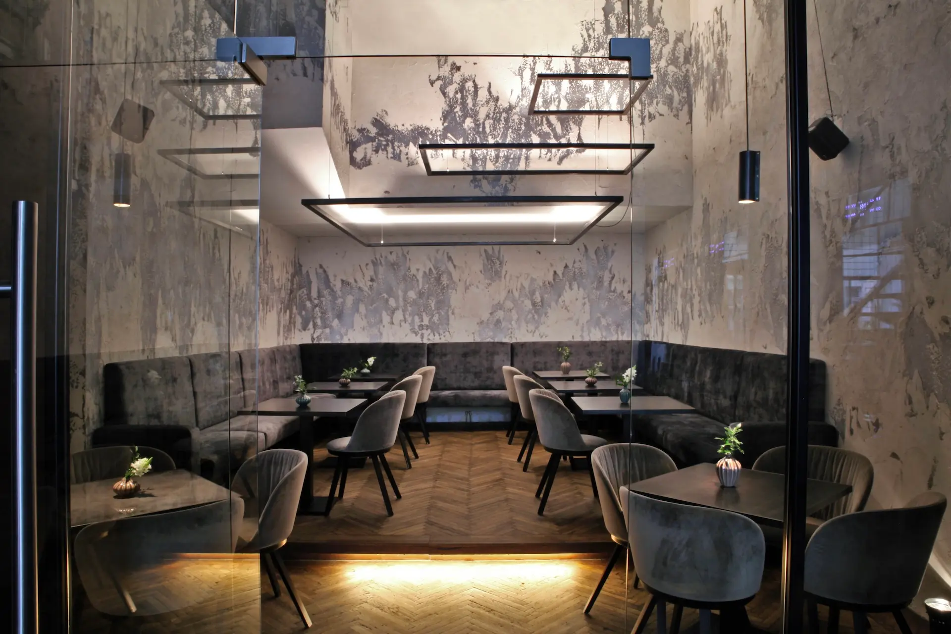Ein modernes, stilvolles Restaurantinterieur mit grauen Stühlen, dunklen Tischen, einer gepolsterten Bank an der Wand, kleinen Pflanzen auf jedem Tisch, gemusterten Wänden und geometrischen Deckenleuchten. Die warme Beleuchtung unterstreicht das schlichte, minimalistische Design.