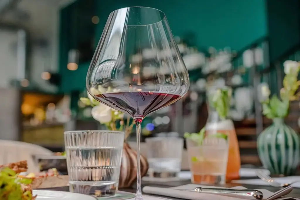 Nahaufnahme eines Rotweinglases auf einem Restauranttisch, umgeben von einem Wasserglas, Utensilien, einer Vase mit Blumen und verschwommenen Getränken und Speisen im Hintergrund.