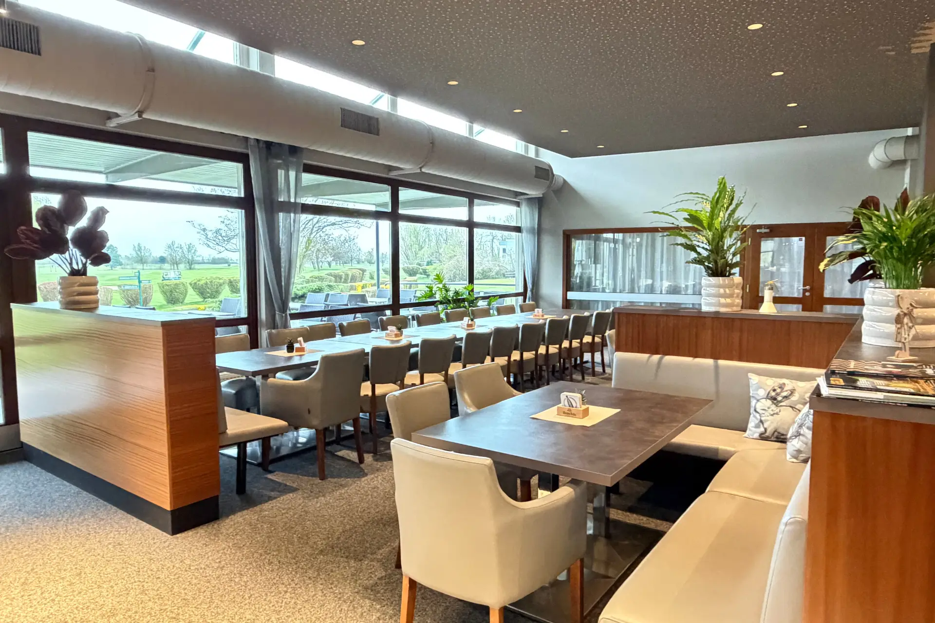 Ein heller Restaurantbereich mit einer Reihe von Tischen und Stühlen entlang einer Wand aus großen Fenstern, die den Blick auf einen Golfplatz freigeben.