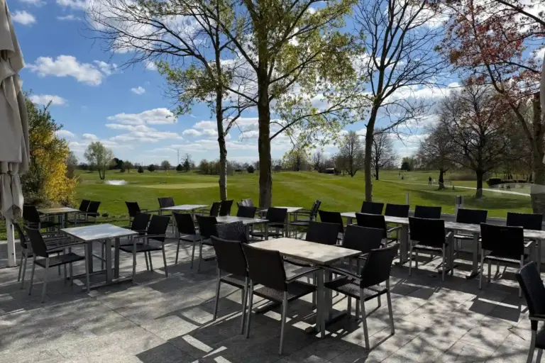 Terrasse im Freien mit mehreren Tischen und Stühlen und Blick auf einen grünen Golfplatz unter blauem Himmel.