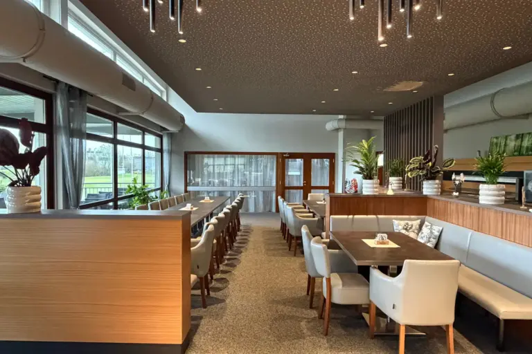 Alleewirt Innenansicht eines modernen Restaurants mit Esstischen, Stühlen und Sitznischen, mit raumhohen Fenstern und einer gemusterten Decke.
