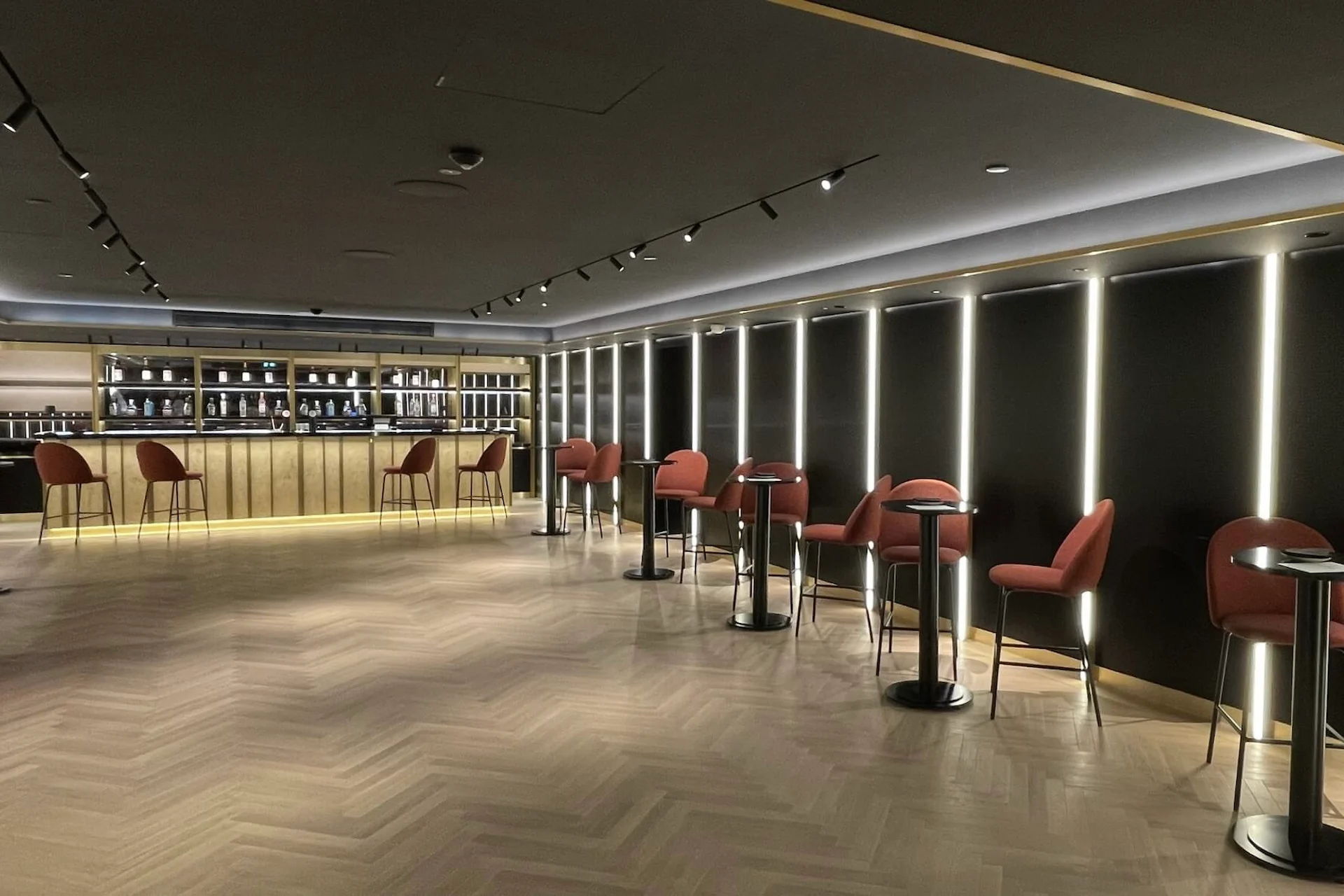 Eine moderne Bar mit einer langen Holztheke, Flaschenregalen und mehreren Stehtischen mit roten, gepolsterten Barhockern. Der Raum ist mit warmen Lichtern gedämpft beleuchtet und hat einen Holzboden mit Fischgrätenmuster.