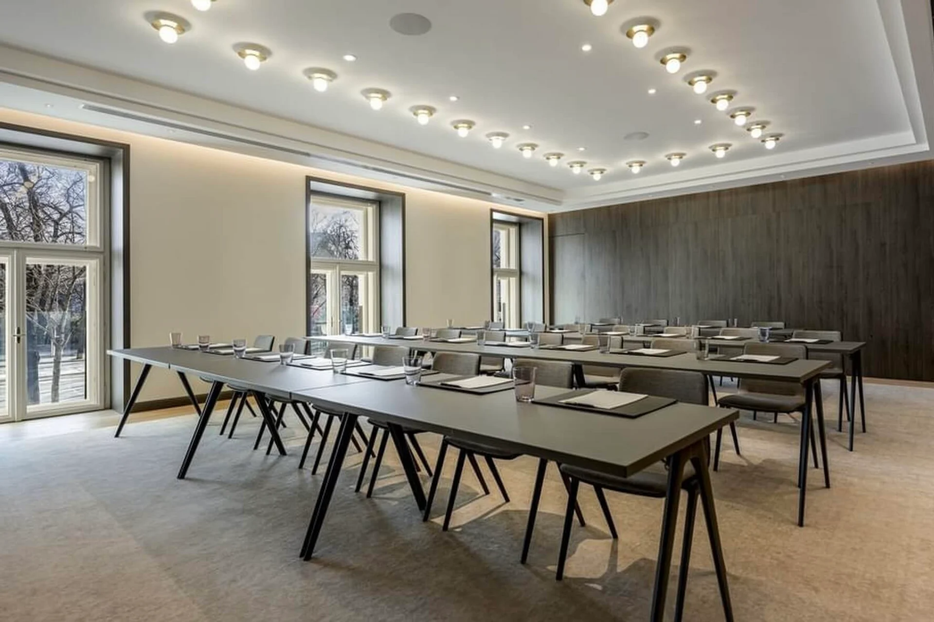 Ein moderner, gut beleuchteter Konferenzraum mit großen Fenstern, mehreren langen, U-förmig angeordneten Tischen, grauen Stühlen und ordentlich angeordneten Notizblöcken und Gläsern auf den Tischen.