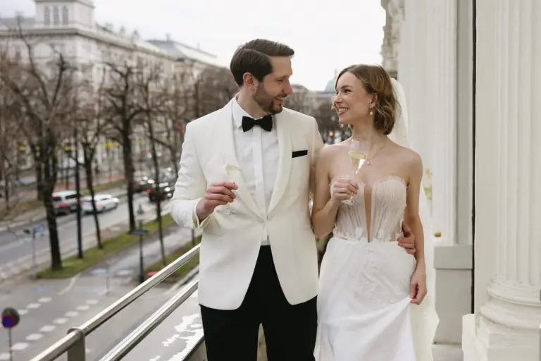 Eine Braut und ein Bräutigam in formeller Hochzeitskleidung stehen auf einem Balkon, lächeln sich an und halten Gläser mit Champagner in der Hand, mit einer Stadtstraße und Gebäuden im Hintergrund.