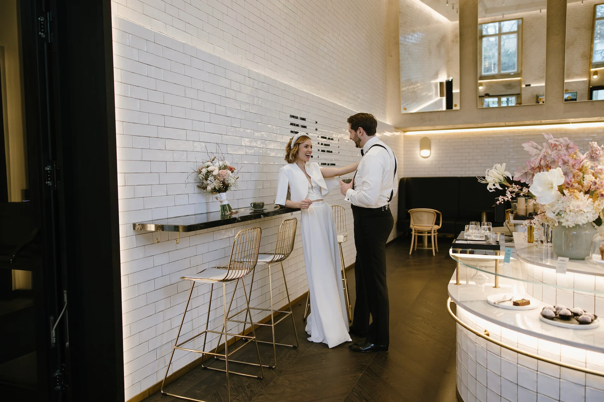 Eine Braut in einem weißen Kleid und ein Bräutigam in Hosenträgern stehen lächelnd neben einem Tresen mit Blumen in einem modernen, weiß gekachelten und gut beleuchteten Restaurant oder Café.