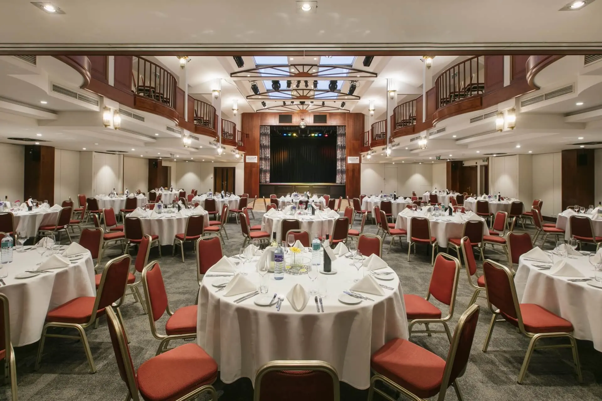 Ein geräumiger Bankettsaal mit runden Tischen mit weißen Tischdecken, roten Stühlen und ordentlichem Geschirr, der für eine Veranstaltung eingerichtet ist. An der Stirnseite befindet sich eine Bühne und an den oberen Wänden sind Balkone angebracht.
