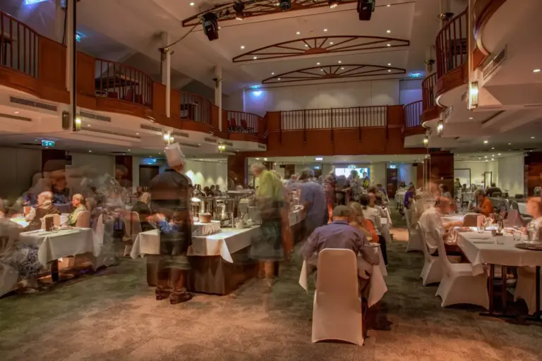 Ein belebtes, elegantes Restaurant mit vielen Gästen an runden Tischen. Der Raum hat hohe Decken, Holzbalkone und eine sanfte Beleuchtung. Einige Figuren erscheinen verschwommen, was auf Bewegung und Aktivität hindeutet.