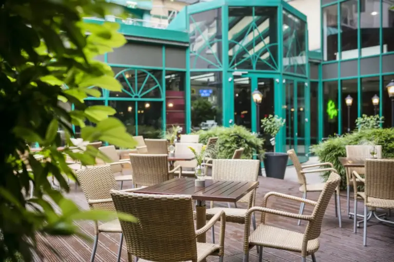 Café-Sitzgruppe im Freien mit Korbstühlen und Holztischen auf einer Terrasse, umgeben von Grünpflanzen. Im Hintergrund ist ein modernes Glasgebäude mit grüner Verkleidung und großen Fenstern zu sehen.