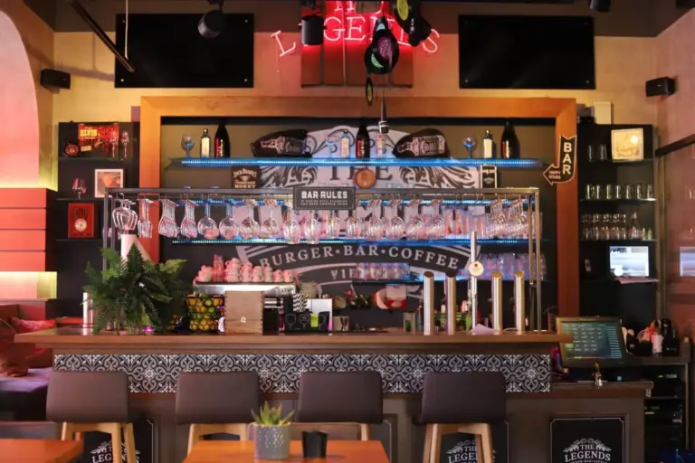 Eine stilvolle Bar mit einem dekorierten Tresen, hängenden Gläsern, Flaschen, einem Neonschild LEGENDS, Pflanzen und Hockern davor. Regale und Bareinrichtungen sind sichtbar und schaffen eine gemütliche, moderne Atmosphäre.