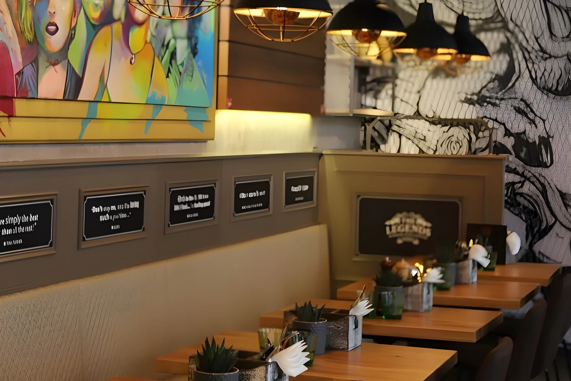 Ein gemütliches Restaurant mit Holztischen, Topfpflanzen und Wandkunst, darunter bunte Gemälde und schwarz-weiße Wandmalereien. Warme Hängelampen erhellen den Sitzbereich, und an der Wand hängen Zitatschilder.