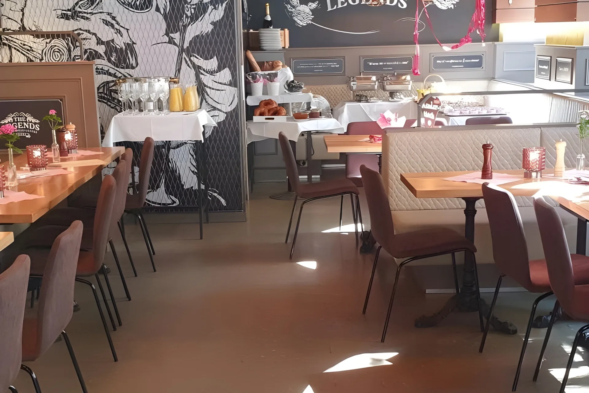 Ein heller Restaurantbereich mit ordentlich aufgestellten Tischen und Stühlen, dekoriert mit Blumen und Kerzen. Im hinteren Bereich stehen Buffet-Tische mit Speisen und Getränken, und an der Wand ist ein Wandgemälde zu sehen.