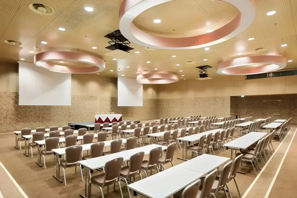 Ein geräumiger Konferenzraum mit Reihen beiger Stühle und weißer Tische vor einer Bühne mit zwei Projektionsflächen und kreisförmigen Deckenleuchten. Die Wände sind hellbraun und haben ein strukturiertes Muster.