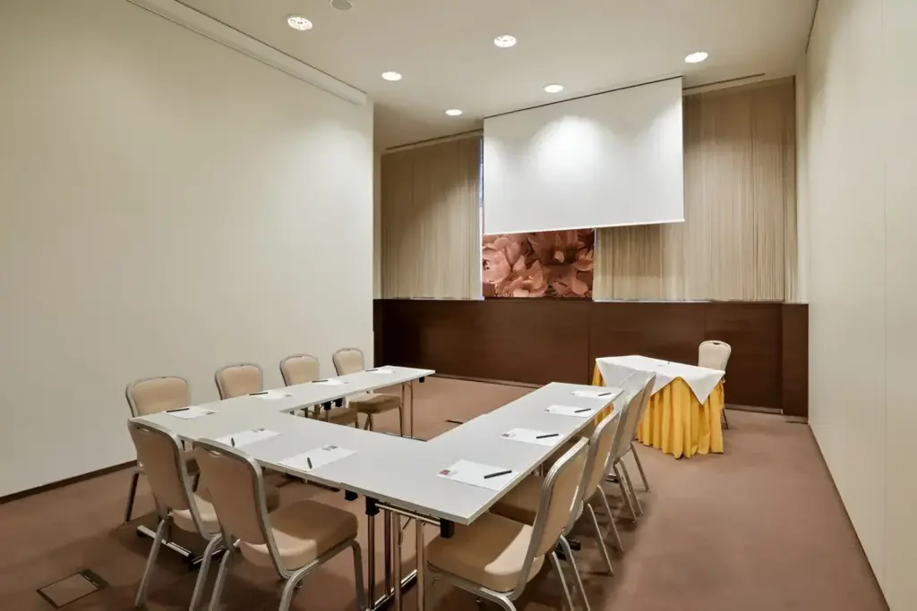 Ein kleiner Konferenzraum mit beigen Wänden, einer U-förmigen Anordnung von Tischen mit Stühlen, Notizblöcken und Stiften. An der Stirnseite steht ein Tisch mit einem gelben Tischtuch und einer eingezogenen Projektionsfläche darüber.