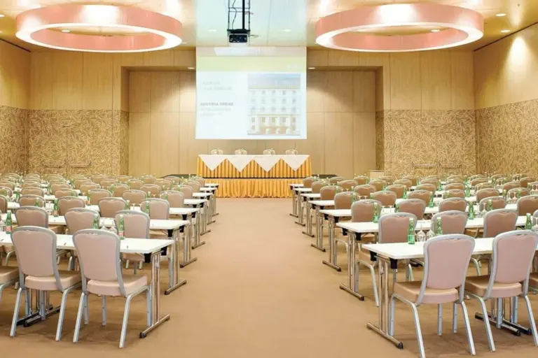 Ein geräumiger Konferenzraum mit Reihen beiger Stühle und Tische vor einer Bühne mit drei Stühlen und einem Tisch mit gelben Röcken. An der Stirnseite ist eine große Projektionsfläche angebracht. Auf jedem Tisch stehen Wasserflaschen.