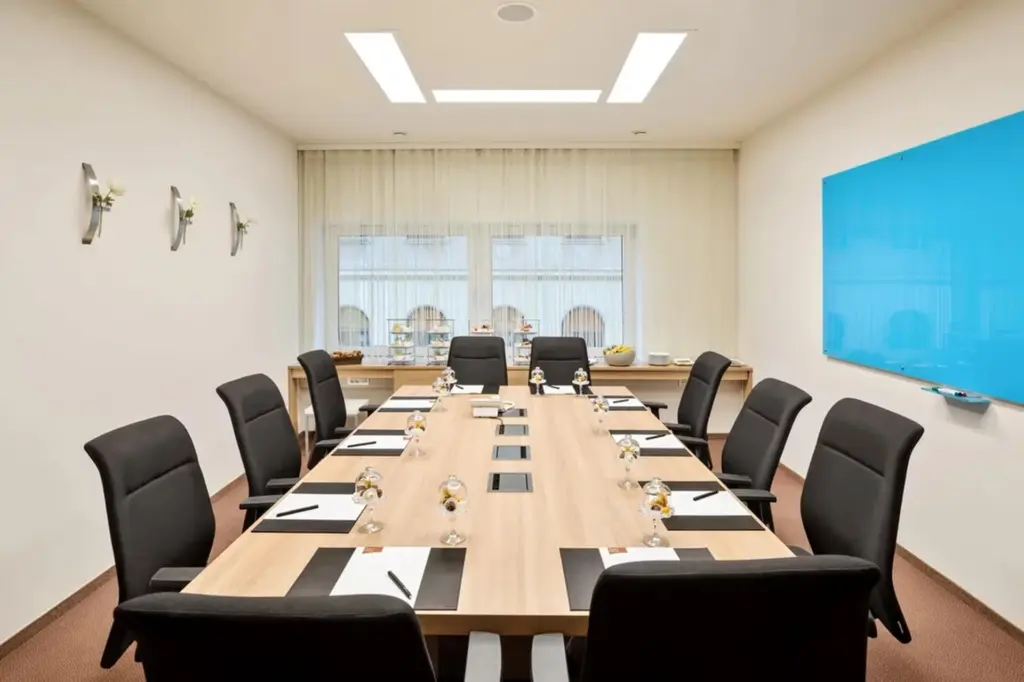 Ein moderner Konferenzraum mit einem langen rechteckigen Tisch, schwarzen Stühlen, Notizblöcken, Stiften, Wassergläsern und kleinen Blumenvasen, mit einer großen blauen Tafel und zwei Fenstern, die Tageslicht hereinlassen.
