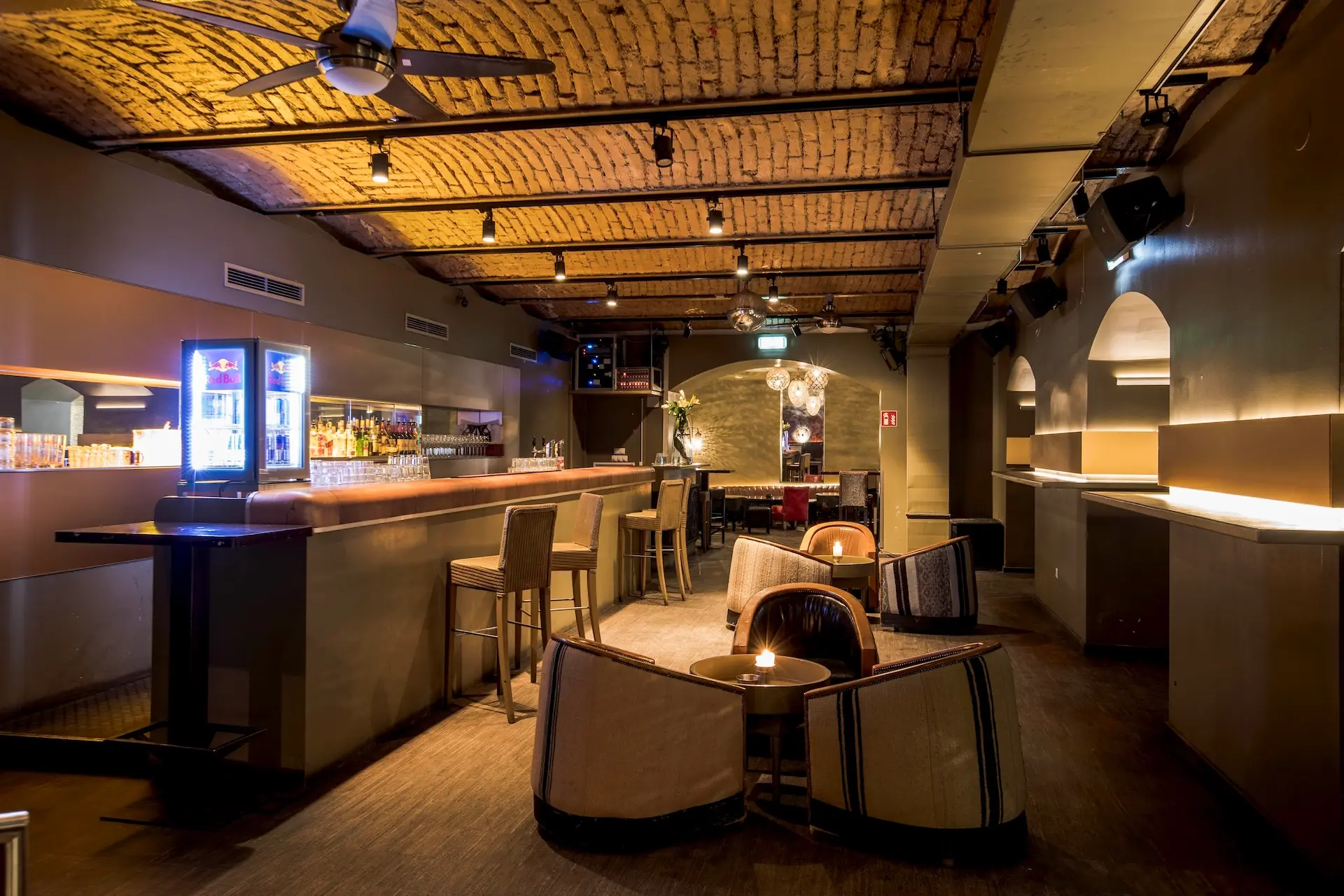 Aux Gazelles Eine stilvolle Bar mit sichtbarer Backsteindecke, moderner Beleuchtung und einem langen Holztresen. Es gibt Barhocker, Loungesessel, kleine Tische mit Kerzen und eine gut gefüllte Bar im Hintergrund. Der Raum ist warm beleuchtet und einladend.