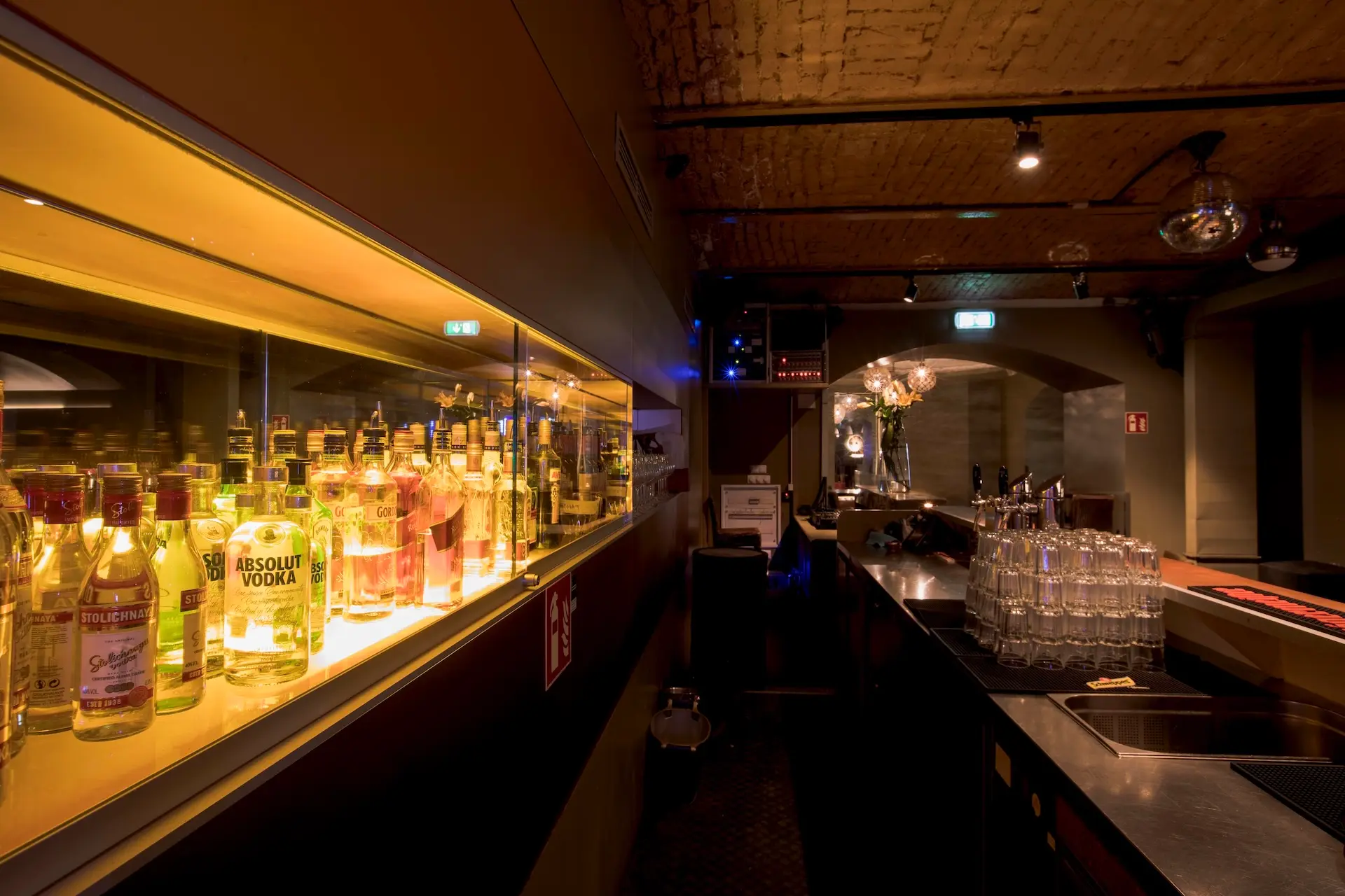 Eine schwach beleuchtete Bar mit Schnapsflaschen in einer Glasvitrine auf der linken Seite und einem Tresen mit Gläsern und Zapfhähnen auf der rechten Seite, unter einer gewölbten Ziegeldecke.