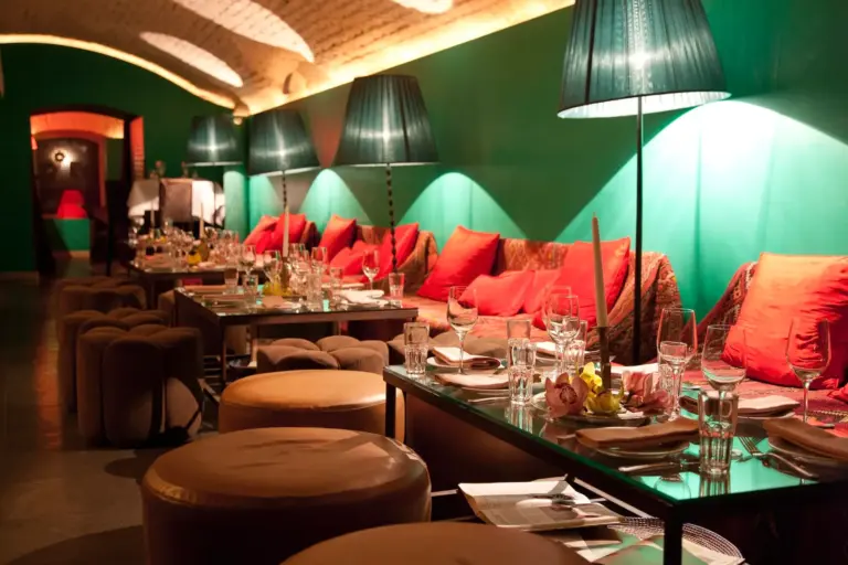 Ein gemütliches Restaurant mit grünen Wänden, roten Polstern und schwacher Beleuchtung durch hohe Lampen. Die Tische sind mit Gläsern und Tellern eingedeckt, und auf einer Seite bieten weiche Hocker und Sofas Platz.