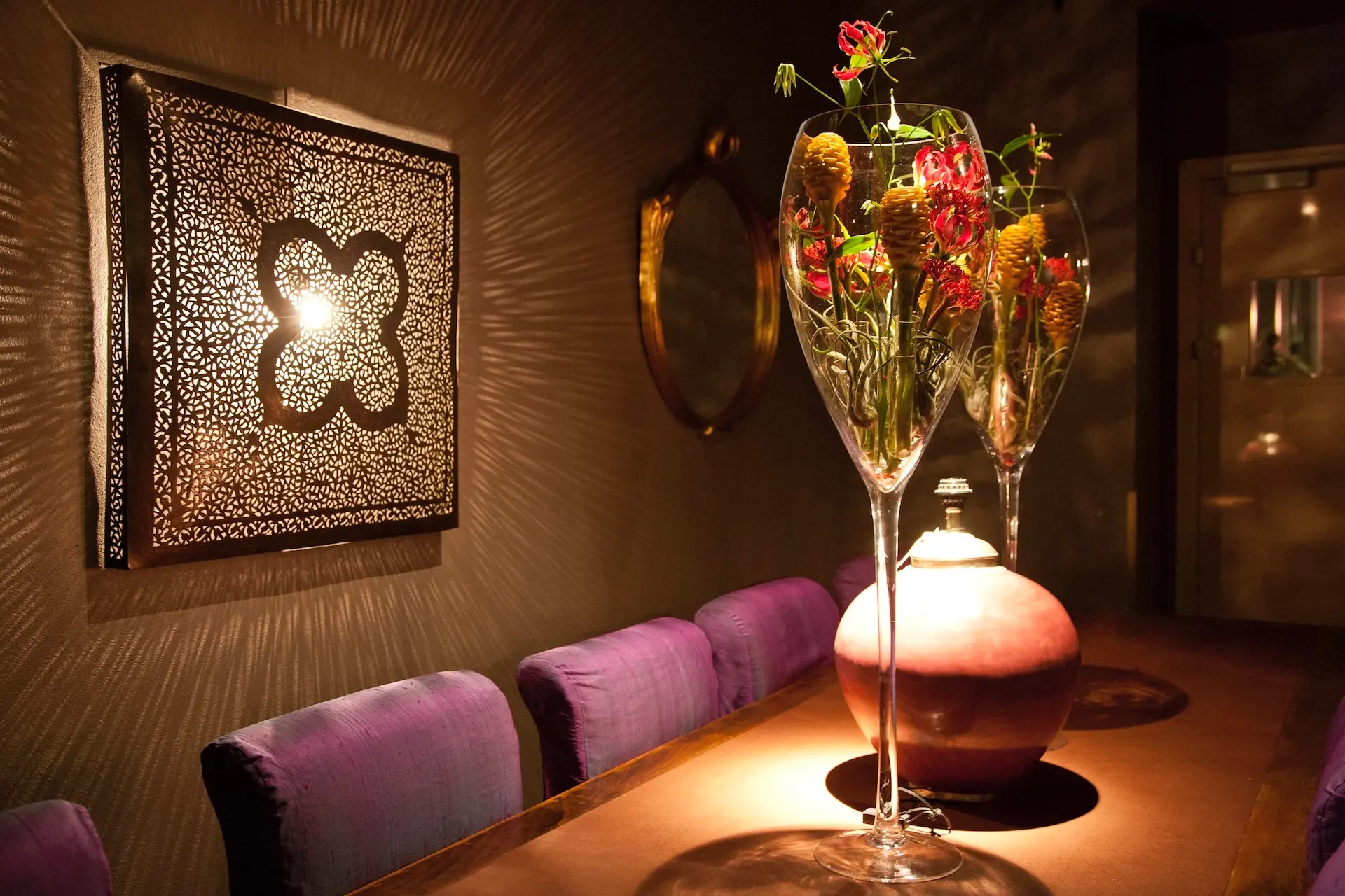 Ein warm beleuchtetes Esszimmer mit einem langen Tisch, violett gepolsterten Stühlen, hohen Glasvasen mit bunten Blumen und einer verzierten Wandleuchte, die kunstvolle Schatten wirft.
