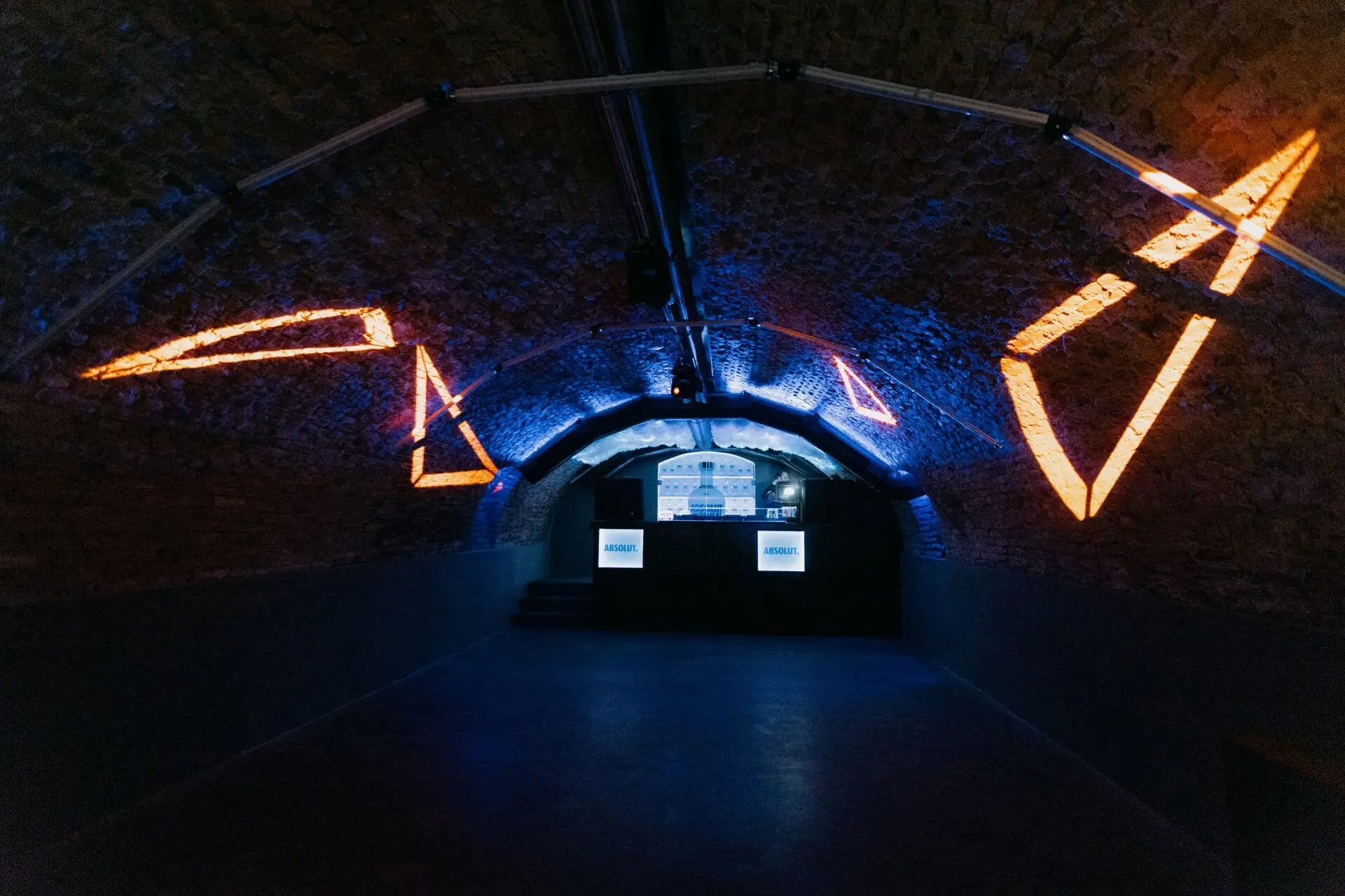 Ein dunkler, gewölbter Backsteintunnel wird mit blauen und orangefarbenen geometrischen Blackshaper-Lichtprojektionen an den Wänden beleuchtet. Am anderen Ende ist eine Bühne mit drei leuchtenden rechteckigen Bildschirmen zu sehen.