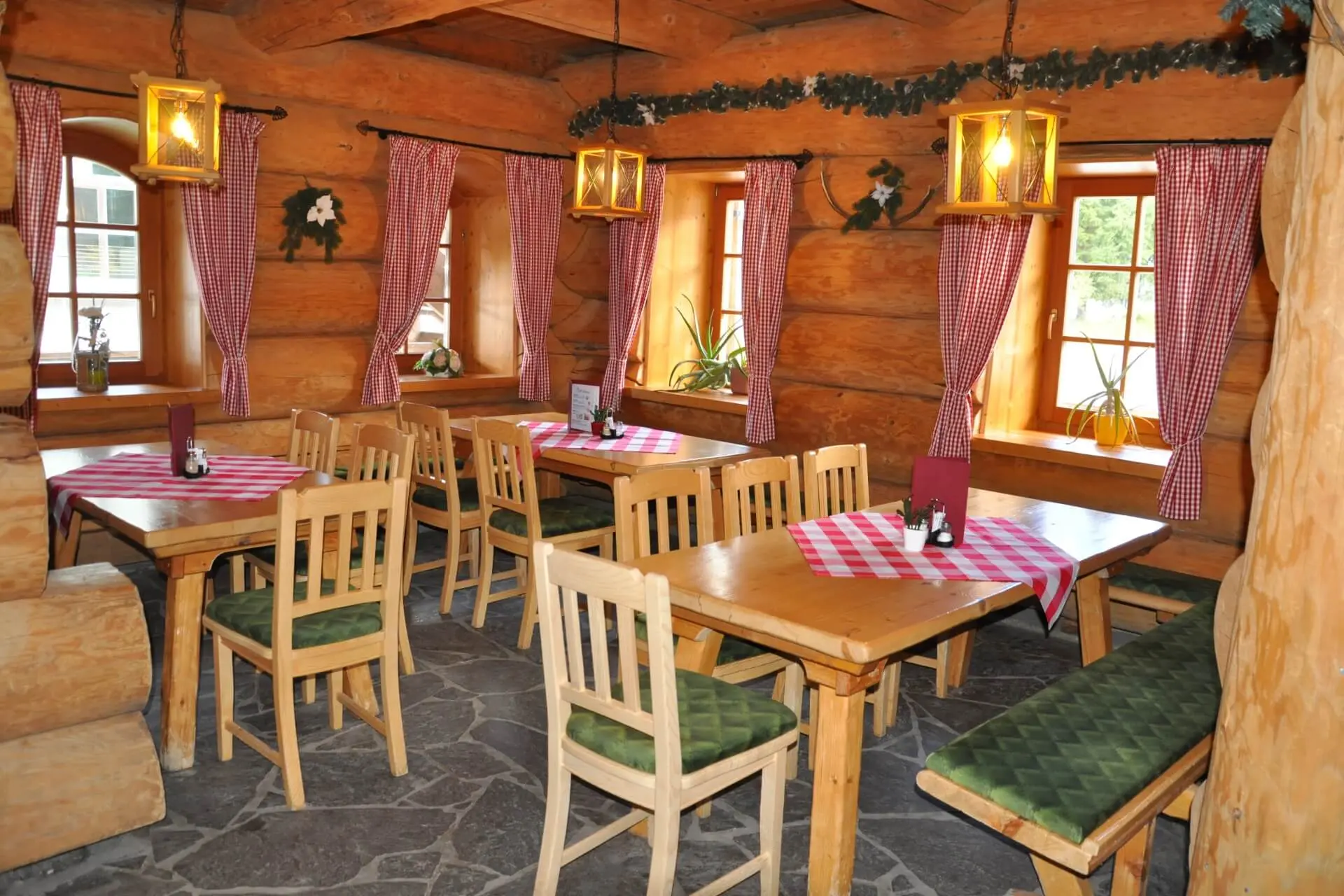 Ein gemütliches Blockhaus-Restaurant mit Holztischen und -stühlen, rot karierten Tischdecken und Vorhängen, Steinboden und von der Decke hängenden Laternen. Durch die Fenster strömt Sonnenlicht.