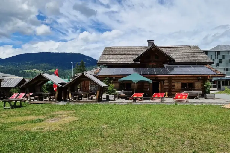 Eine rustikale Holzhütte mit Sonnenkollektoren auf dem Dach steht neben Liegestühlen und Sitzgelegenheiten im Freien unter Sonnenschirmen, umgeben von grünem Gras und Bergen unter einem teilweise bewölkten Himmel.