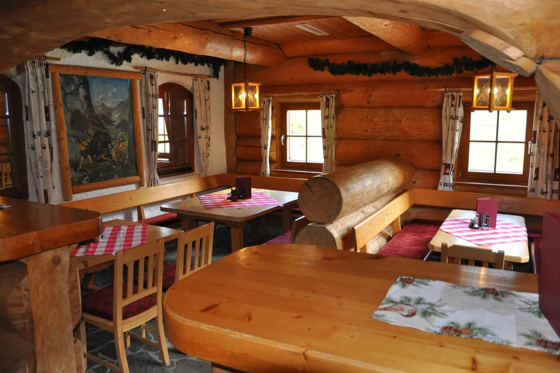 Ein gemütliches, rustikales Blockhaus-Restaurant mit Holztischen und -bänken, rotkarierten Tischdecken, warmer Beleuchtung und Walddekoration, einschließlich gerahmter Kunst und Girlanden über den Fenstern.