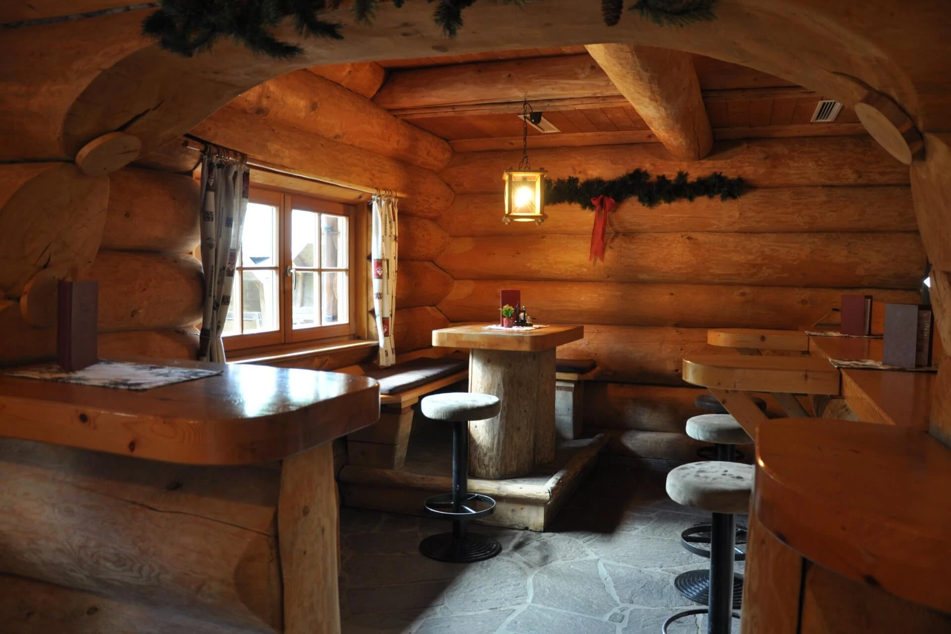 Eine gemütliche Blockhütte mit Holztischen und -hockern, einem Fenster mit Vorhängen, die natürliches Licht hereinlassen, Steinböden und festlichem Grün, das von der Decke hängt.